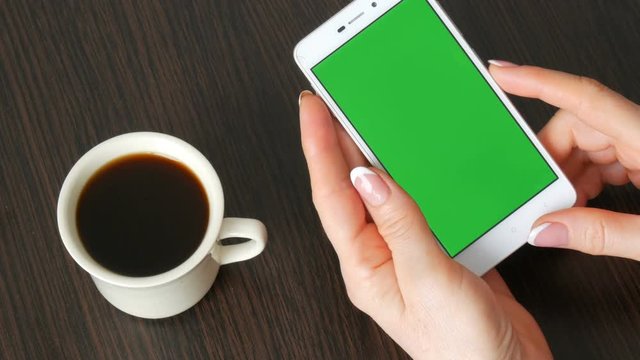 Female hands with beautiful French manicure take a white smartphone with Green Screen near white cup of coffee. Using Smartphone,Holding Smartphone with Green Screen