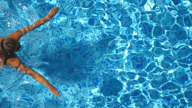 Top view of unrecognizable young girl in red bikini swimming across the pool of hotel. Beautiful woman floating in basin and relaxing on sunny day. Concept of summer vacation or holiday. Close up
