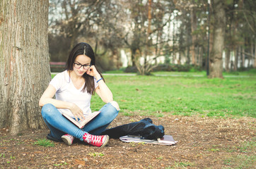 Education concept. Beautiful young school or college girl with long hair and glasses sitting on the ground in the park reading the books and study for exam