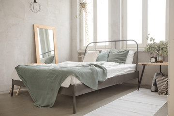 Bed with green coverlet stands in bright bedroom with large French windows mirror and small bedside lamp with a plant and alarm clock. Well decorated bedroom in soft lights