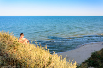 A young man in a red shirt sits on a hill in the background of the sea