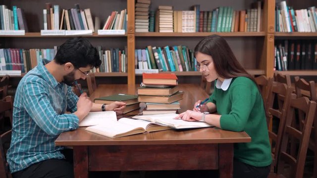 Two smart students working on home assignment, reading books attentively, sitting at square library table next to the bookshelves, curious girl pointing at the text in boy's book