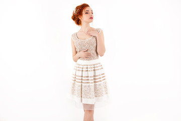 young girl with red hair in a beige lace dress standing like princess queen with hand on her collarbone on a white wall background