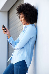 Side of relaxed young african woman using a mobile phone leaning to a wall outdoors