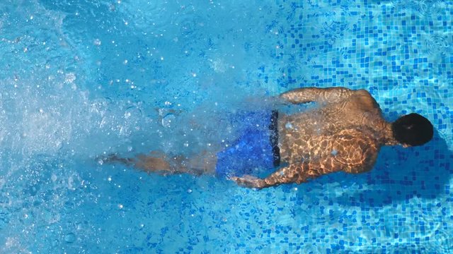 Top view of unrecognizable sporty man jumping in clear blue water of pool. Young muscular guy floating across the basin of hotel on sunny day. Concept of summer vacation or holiday. Close up