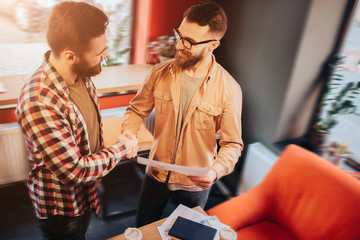 Two hipsters that wear casual clothes standing in a slall cafe and shaking their hands.One of them is holdnig a document in his hand. Both of them are satisfied with their collaboration.