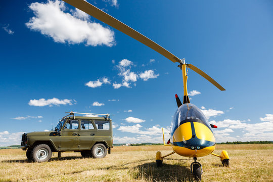A small yellow light-engine helicopter stands next to a Russian green military off-road car in the middle of a field with grass against the background of a bright blue sky on a summer clear sunny day