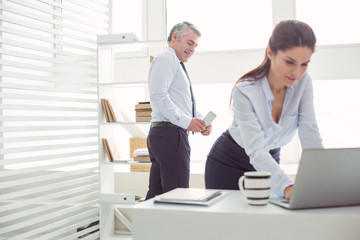 Obraz premium Gender discrimination. Unpleasant cheerful adult man standing behind his colleague and taking a photo of her while using his smartphone