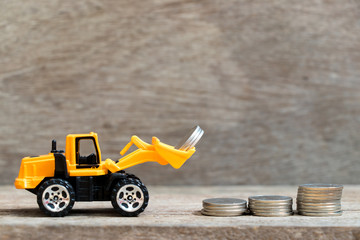 Fototapeta premium Toy bulldozer hold group of coin to fulfill up trend of stacked coin on wood background