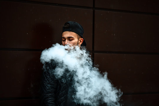Vape man. Portrait of a handsome young white guy in a modern black cap vaping and letting off puffs of steam from an electronic cigarette opposite the futuristic urban background.