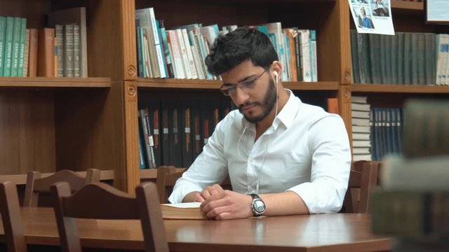 Good-looking asian man reading book, serious guy with black wavy hair wearing white headphones and glasses, sitting at the brown table next to the library bookshelves