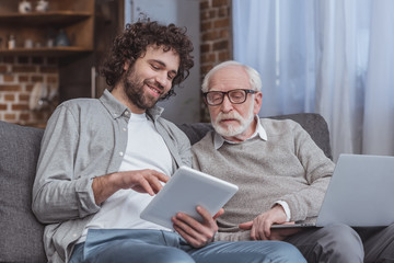 adult son and senior father using tablet and laptop at home