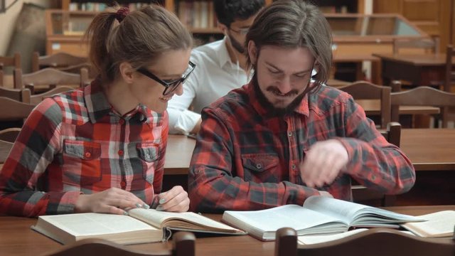 Two happy friends reading in large light library, smart bearded boy looking through the pages, bright girl in glasses talking to the mate, wearing stylish checked shirts