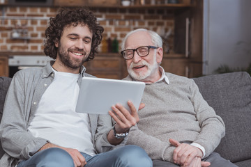 smiling adult son showing something on tablet to senior father at home