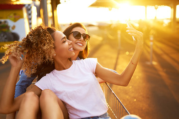 Selfie time! Two beautiful girls having fun on shopping trolleys. Multiethnic young people racing...