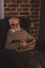 Obraz premium handsome grey hair man reading book in armchair at home