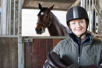 Portrait Of Female Owner Holding Saddle In Stable With Horse - Powered by Adobe