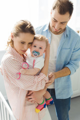 Mother holding infant daughter with baby dummy and father standing near