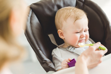 Infant daughter in baby chair rejecting from food that mother giving to her