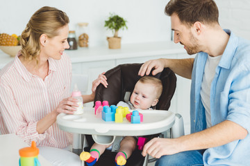 Parents with infant daughter sitting in baby chair with plastic blocks and feeding bottle