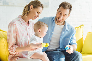 Young parents sitting on sofa with infant daughter holding plastic blocks