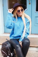 Obraz na płótnie Canvas Close up fashion portrait of a happy young woman sitting on the city stairs.smiling young blonde traveler in hat resting while sitting on staircase outdoors.Wearing blue sweater,jeans and backpack.