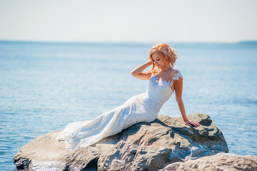 Fototapeta na wymiar Beautiful, slender bride in a silhouette wedding dress posing on the stone beach on a Sunny day. Blue sky, bright sun.