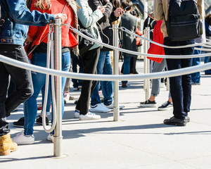 People standing in line between stainless steel queue poles with grey ropes, at the entrance of a tourist site, leaning on the poles or watching their smartphones.