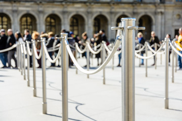 Stainless steel poles linked by grey ropes for queue control at the entrance of a tourist site, with drop shadows on a light ground and blurry people queueing up in the background.
