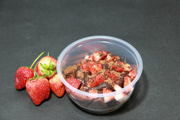 Home made cake and chocolate lava, topping by Strawberries cut into pieces with cocoa powder in the round plastic cup. And Strawberry on the black floor.