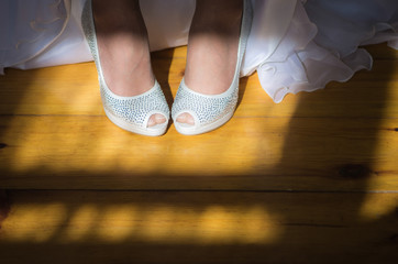 Closeup top view of female legs in white and silver shiny high heels. Beautiful young bride in long gorgeous dress indoors. Shadow of window on floor. Horizontal color photography.