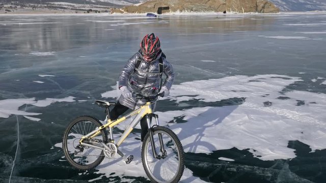 Woman is walking beside bicycle on the ice. The girl is dressed in a silvery down jacket, backpack and helmet. Ice of the frozen Lake Baikal. The tires on the bicycle are covered with special spikes