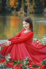 Девочка в красном платье с рябиной сидит на деревянном пирсе на озере