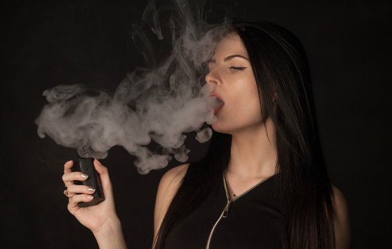 Young beautiful brunette woman smoking, vaping e-cigarette with smoke on black background.