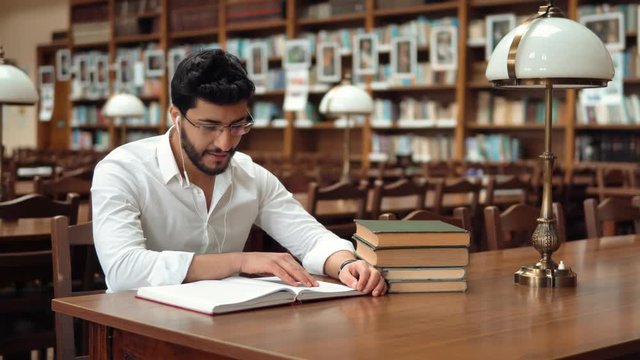 Good-looking slim man wearing white headphones while reading, smiling student relaxing with book and music at brown wooden table in nice big library