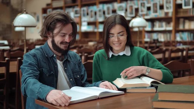 Two intellectual teenagers are reading together, discussing information as turning the pages, lovely long-haired girl wearing green sweater and stylish glasses, bearded boy - blue shirt and white t