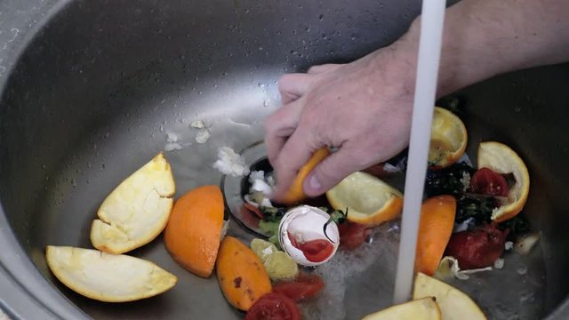 Kitchen sink with a shredder of household waste. The sink is well-used. A stream of water from the tap. Eggshells, oranges and rotten tomatoes.
