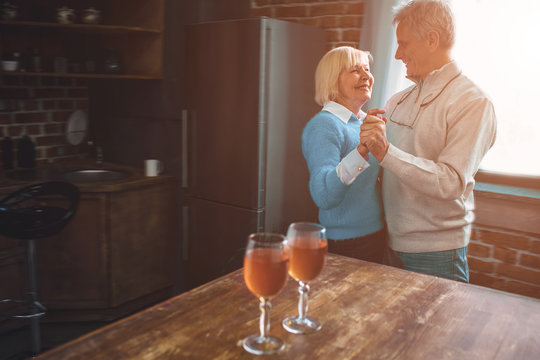 Nice and warm picture of old couple dancing together in the kitchen. They have left two full wineglasses of wine on the table. They are enjoying company of each other.