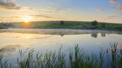 . Sunny summer morning.Foggy landscape with river.Beautiful clouds in sky.Fog over the water.Warm sunlight at sunrise.River Krasivaya in Tula region,Russia. 