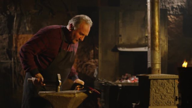 Blacksmith brings dripping hot metal from furnace and he beats a piece of hot metal with a hammer on an anvil. Shot on RED EPIC-W Helium Cinema Camera in Slow Motion.