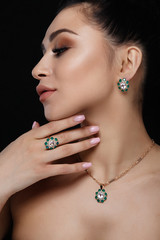 Charming model with dark hair shows rich golden earrings, necklace, and ring with green precious stones
