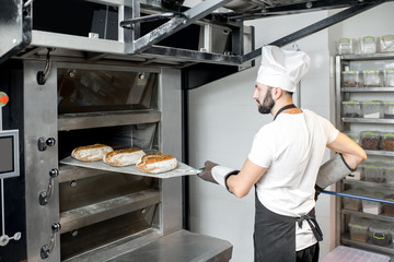 Baker taking off baked breads with shovel from the professional oven at the manufacturing