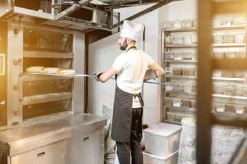 Baker taking off baked breads with shovel from the professional oven at the manufacturing
