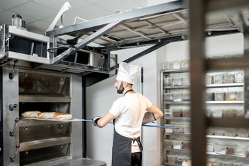 Baker taking off baked breads with shovel from the professional oven at the manufacturing