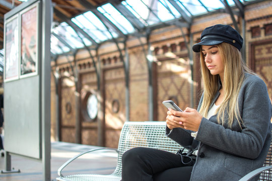 Young woman wearing coat and hat using phone while waiting for train at railway platform.