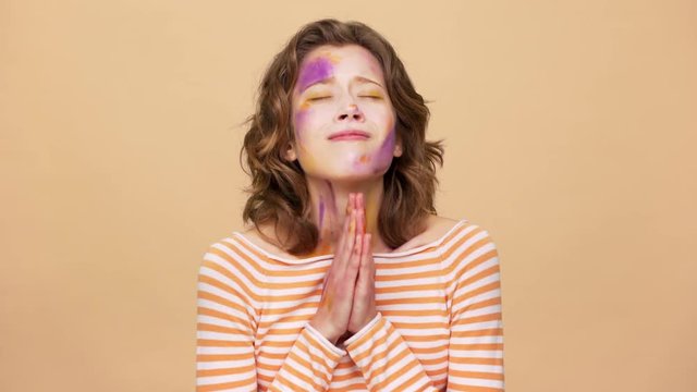 Portrait of dreaming artistic woman wearing multicolored makeup stains on face and praying god please holding palms together, isolated over beige background. Concept of emotions