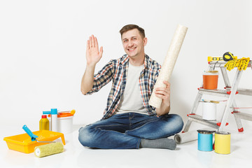 Young smiling man sitting on floor with waving hand for greeting. Instruments for renovation apartment isolated on white background. Wallpaper, gluing accessories, painting tools. Repair home concept.