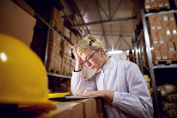 Young worried dedicated female employee in facility storage area is checking paper sheets leaned on a stack of boxes in factory storage room.