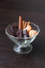 Chocolate with cinnamon sticks and tangerines in a transparent dessert plate on a dark table