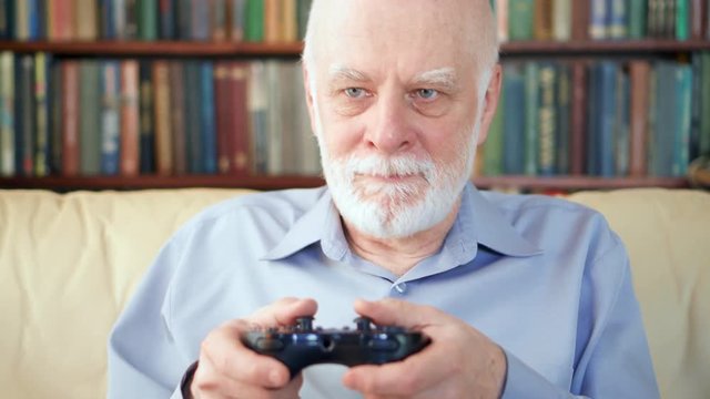 Senior man gamer playing video games at home. Addicted senior player with remote controller of game console. Cybersport for active modern elderly people. Bookcase bookshelves in the background